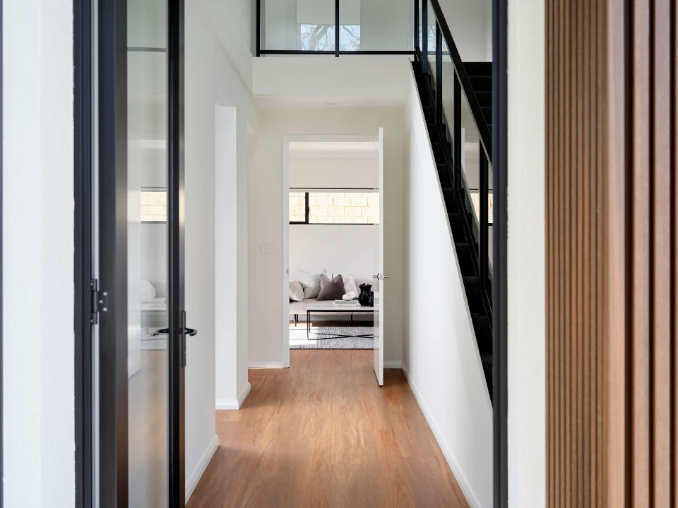 Modern home hallway with wooden flooring, black staircase, and bright natural light from open room.