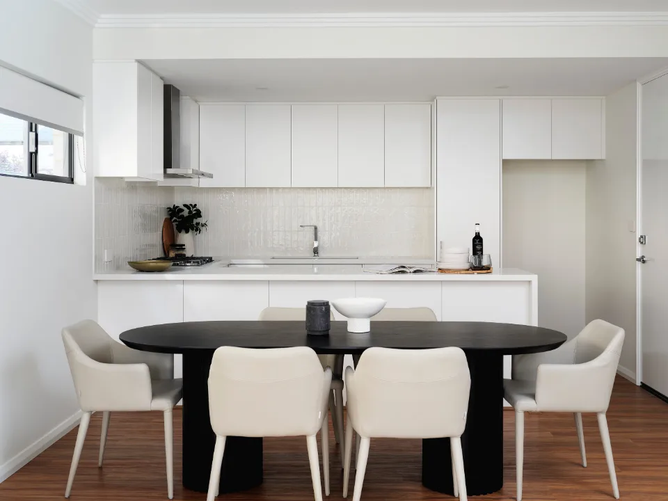 Modern white kitchen with sleek cabinets, wooden floor, and elegant dining table set with a black oval table and six chairs.
