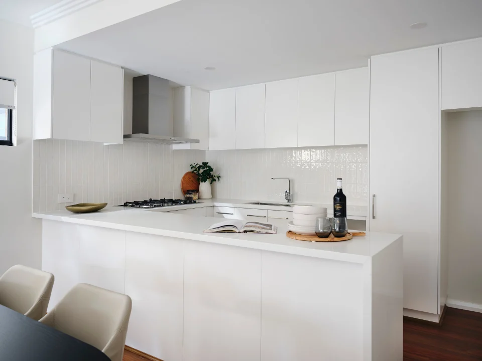 Modern white kitchen with sleek cabinets, island, and cooktop, featuring minimalist design and wooden floor.