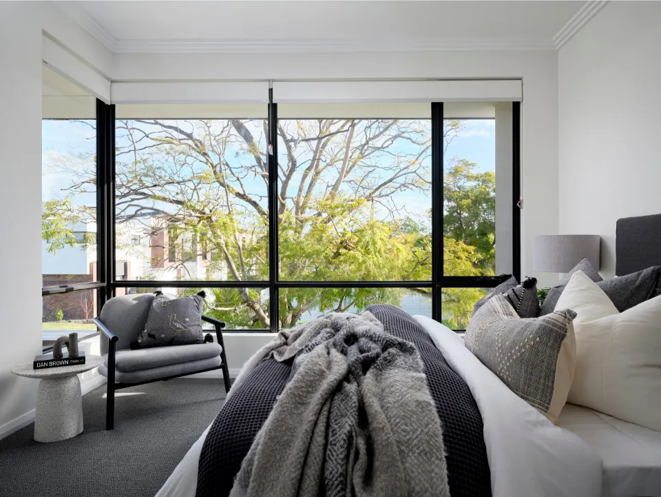 Modern bedroom with large window view of trees in natural light. Cozy bed with gray and white pillows and throw blanket.