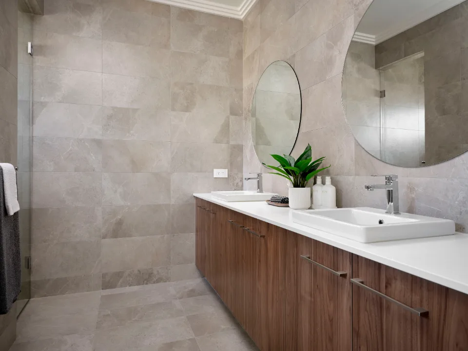 Modern bathroom with dual sinks, wood cabinets, large mirrors, and potted plant on a tiled wall.