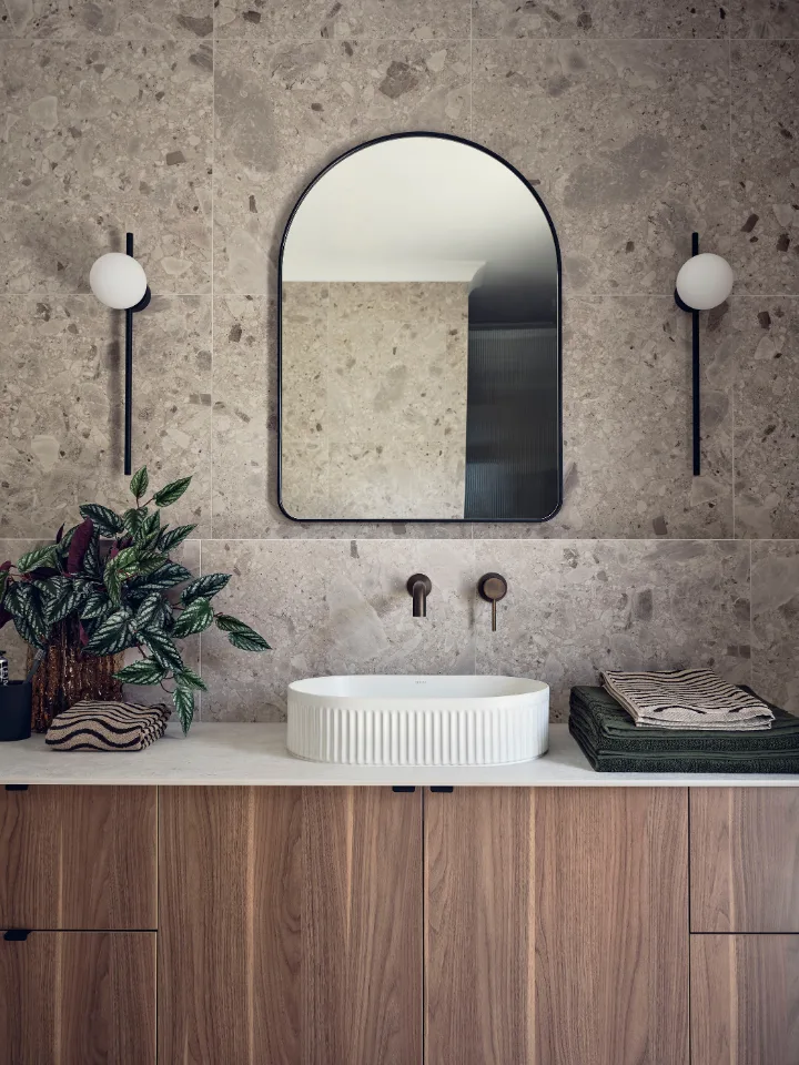 Modern bathroom with stone wall, oval mirror, wooden vanity, and plant decor.