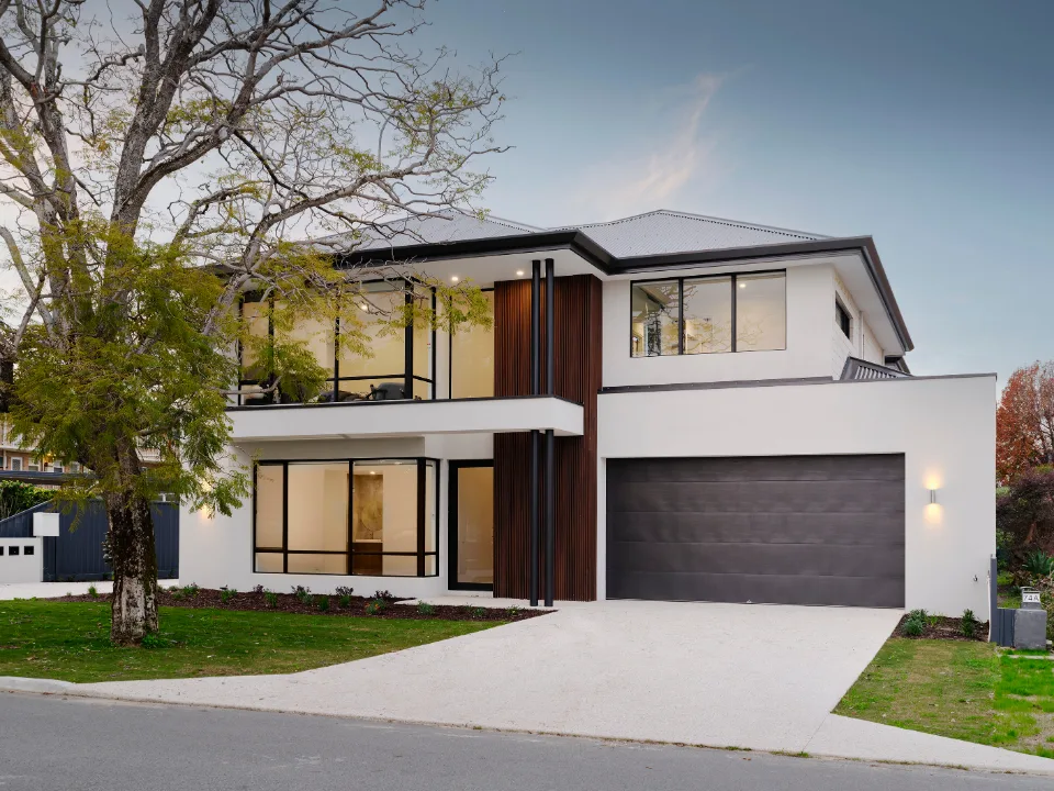 Modern two-story house with large windows, garage, and tree-lined yard at dusk.