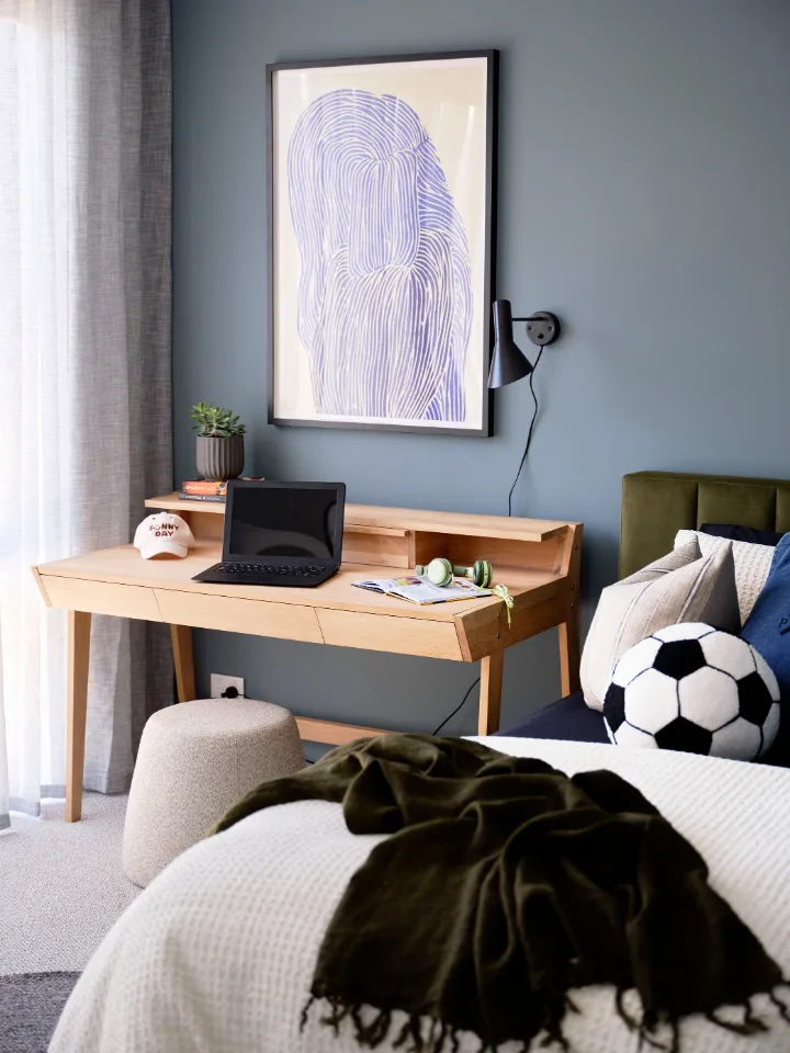 Stylish bedroom with wood desk, laptop, and artwork on blue wall. Cozy bed features a soccer ball pillow and a dark blanket.