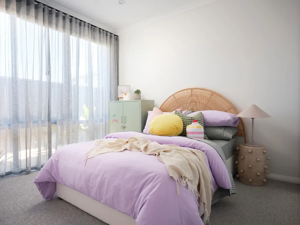 Cozy bedroom with lavender bedding, wicker headboard, and sunlight streaming through sheer curtains.
