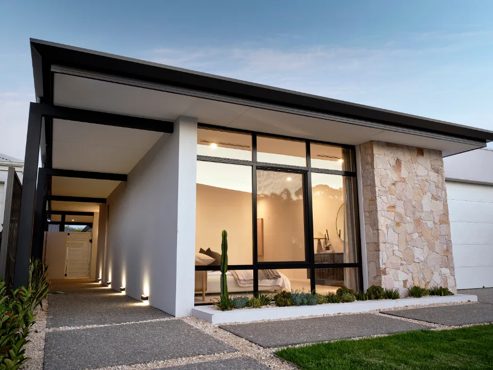 Modern house exterior with large windows and stone facade, featuring a lit pathway and a small garden.
