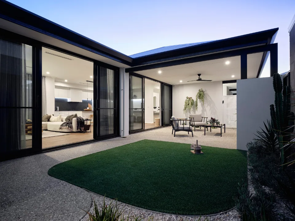 Modern patio with seating area, greenery, and sliding glass doors leading to an illuminated living room at dusk.