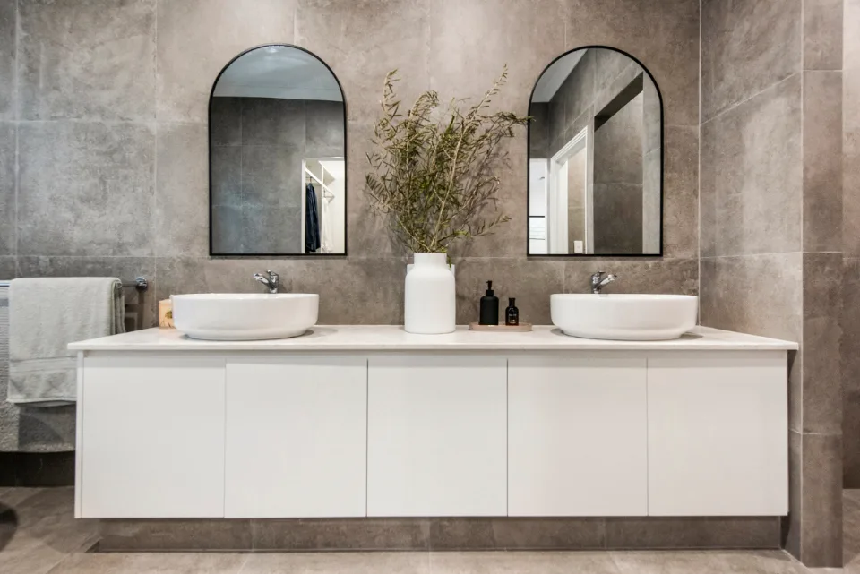 Modern bathroom with double sinks, arched mirrors, and a vase on a marble countertop.