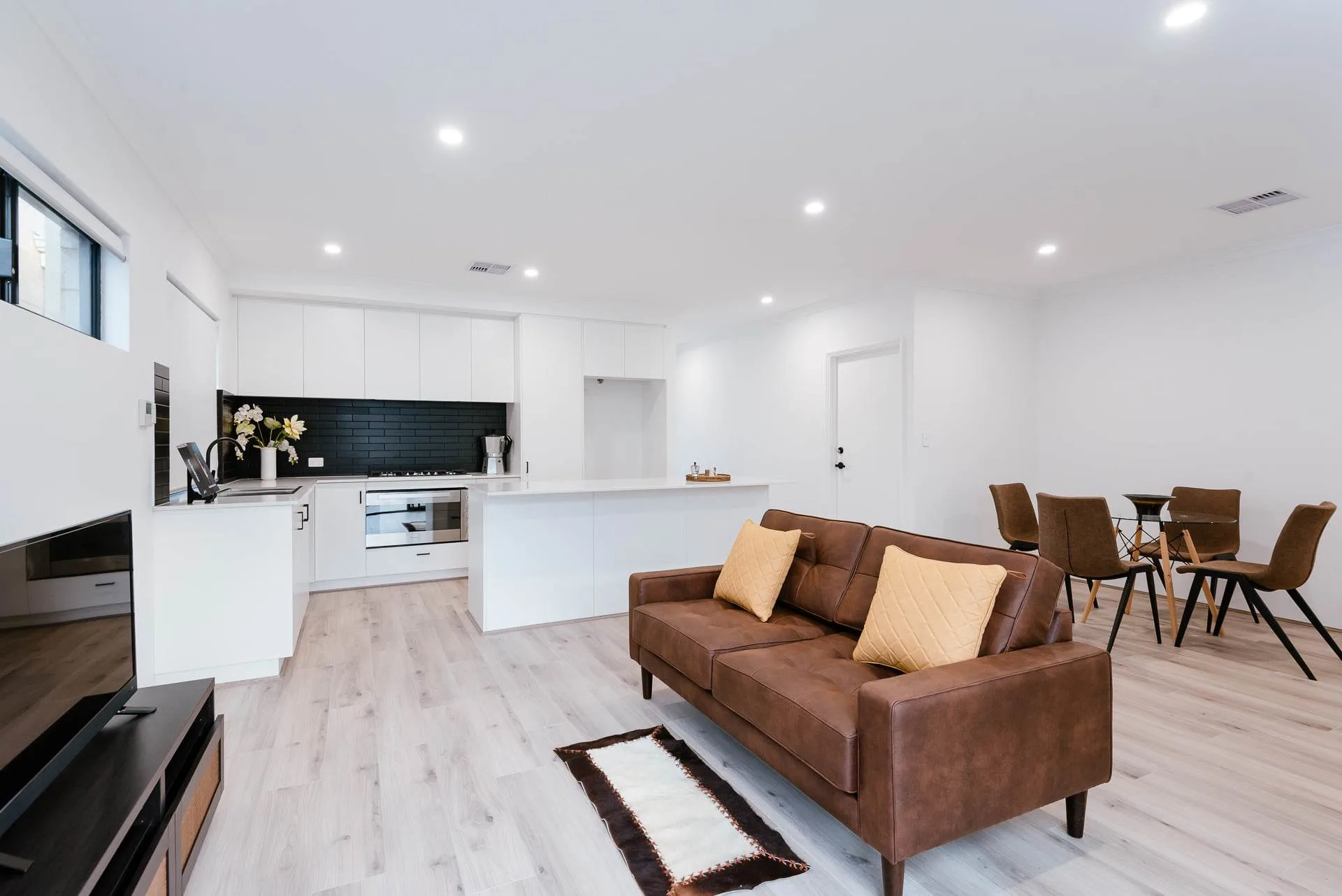 Modern open-plan kitchen and living area with brown sofa and sleek white cabinetry.