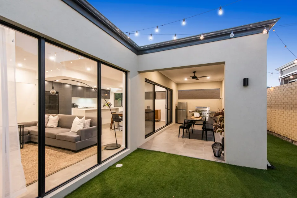 Modern patio with outdoor dining, string lights, and large glass doors leading to stylish living room at dusk.