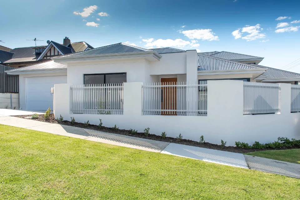 Modern white suburban house with metal fence and lush green lawn under a blue sky.