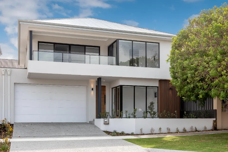 Modern two-story house with large windows and garage, surrounded by greenery and a clear blue sky.