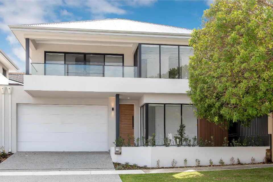 Modern two-story house with large windows and garage, surrounded by lush greenery under a blue sky.
