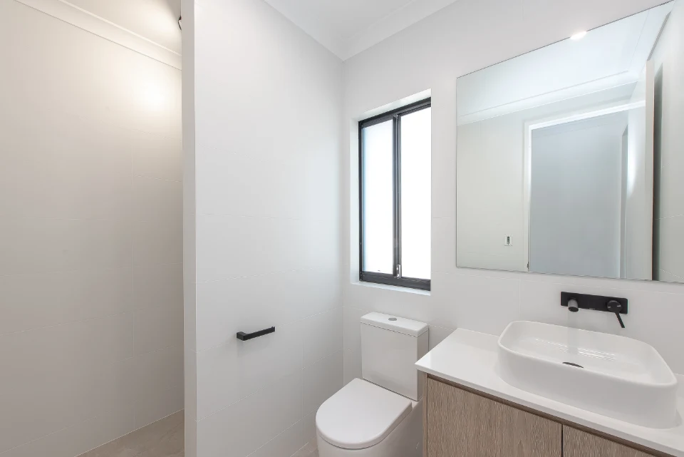 Minimalist white bathroom with modern fixtures, large mirror, and natural light from a window.