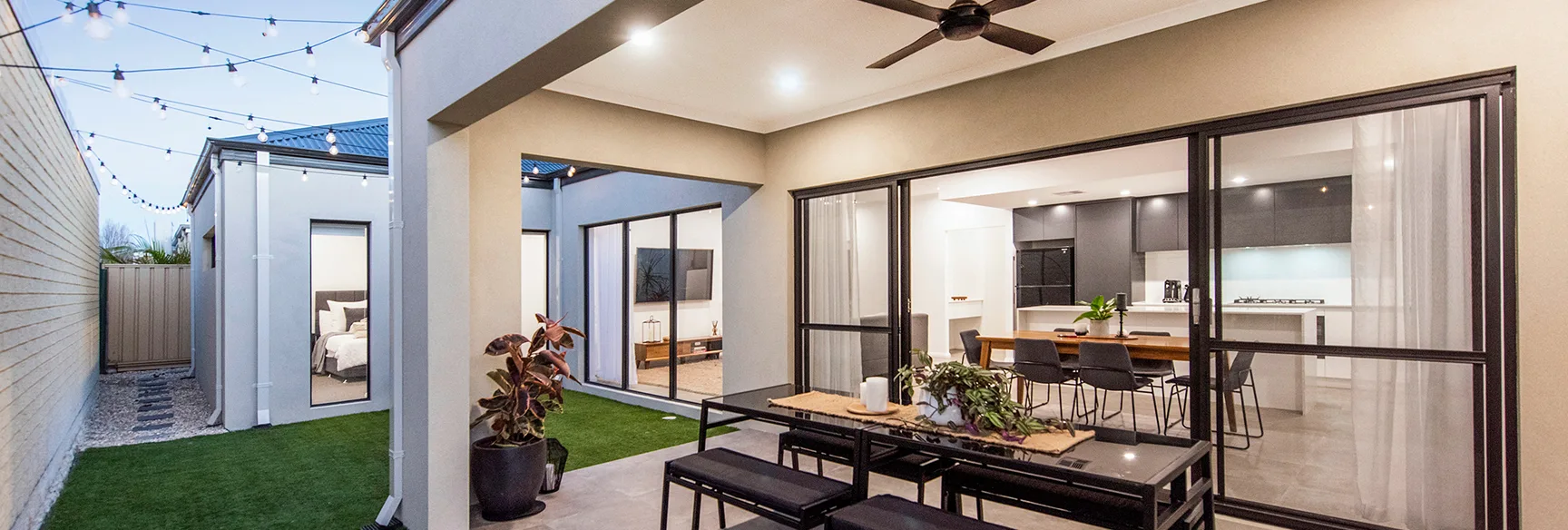 Modern patio with table, string lights, and view into a sleek kitchen and living room through large glass doors.
